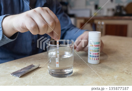 A man in the kitchen tests tap water quality with a test strip. Home testing for drinking water safety, pH, chlorine, and chemical contaminants. A man in the kitchen tests tap water quality with a test strip. Home testing for drinking water safety, pH, chlorine, and chemical contaminants. 135877187