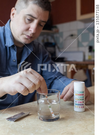 A man in the kitchen tests tap water quality with a test strip. Home testing for drinking water safety, pH, chlorine, and chemical contaminants. A man in the kitchen tests tap water quality with a test strip. Home testing for drinking water safety, pH, chlorine, and chemical contaminants. 135877188