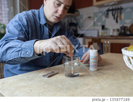 A man in the kitchen tests tap water quality with a test strip. Home testing for drinking water safety, pH, chlorine, and chemical contaminants. 135877189