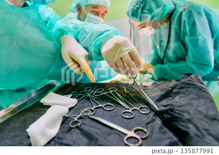 Surgical Team In Green Scrubs Performing Operation With Instruments On Tray In The OR 135877991