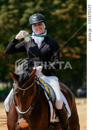 A female hijab equestrian athlete on horseback is holding a gold medal after winning a dressage competition. This image represents achievement, success, and dedication in sports, showcasing the A female hijab equestrian athlete on horseback is holding a gold medal after winning a dressage competition. This image represents achievement, success, and dedication in sports, showcasing the 135878125