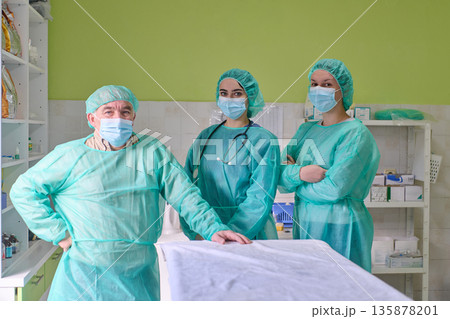 Medical Team in Green Surgical Gowns and Masks Stands by Patient in Operating Room Medical Team in Green Surgical Gowns and Masks Stands by Patient in Operating Room 135878201