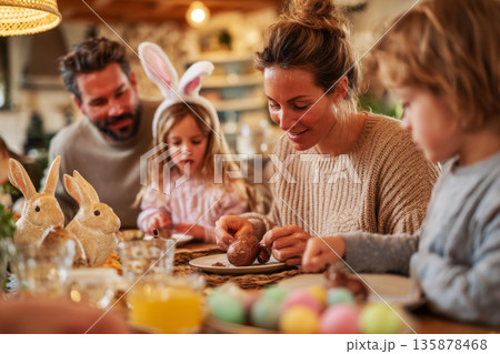 Family enjoys time together at a decorated table for Easter. They exchange chocolate bunnies and colorful eggs while smiling and sharing laughter. Children express joy and excitement 135878468