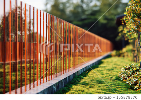 A semi-transparent glass fence marks the boundary of a private garden. The setting is a modern suburban neighborhood with greenery and a home in the background 135878570