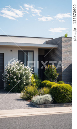 A modern suburban house with a stylish grey brick boundary wall stands amid lush landscaping under a clear sky. The scene captures the tranquility of a residential area 135878600