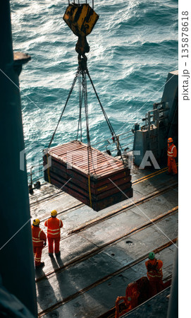 Crew in work clothes uses a crane to lift heavy containers filled with seabed minerals. The wet deck reflects sunlight as the sun sets in the background 135878618