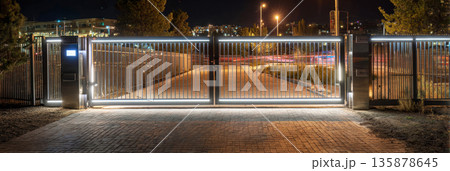 A high-tech gate featuring a fingerprint scanner and solar panels stands elegantly in a well-lit suburban area at night. The combination of metal and wood offers a sleek appearance, banner A high-tech gate featuring a fingerprint scanner and solar panels stands elegantly in a well-lit suburban area at night. The combination of metal and wood offers a sleek appearance, banner 135878645