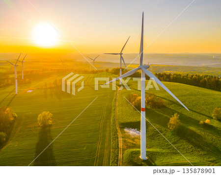 Wind turbines are rotating in a field during sunset. The sun casts a bright light over the landscape. Trees are visible nearby as shadows stretch across the ground. Wind turbines are rotating in a field during sunset. The sun casts a bright light over the landscape. Trees are visible nearby as shadows stretch across the ground. 135878901