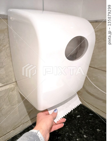 A woman's hand pulls out a white paper towel from a wall-mounted dispenser. A woman's hand pulls out a white paper towel from a wall-mounted dispenser. 135879299
