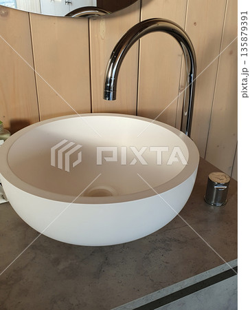 A stylish, modern bowl sink with a smooth matte white finish sits on a concrete countertop. A chrome faucet with an arched spout complements the minimalist, wood-paneled bathroom interior. A stylish, modern bowl sink with a smooth matte white finish sits on a concrete countertop. A chrome faucet with an arched spout complements the minimalist, wood-paneled bathroom interior. 135879391
