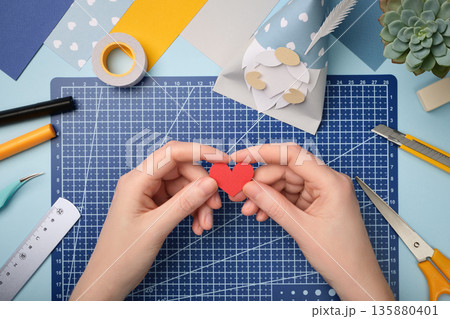 Making a gnome in love craft. Hands are holding a red heart cut out of paper. Creative process. Making a gnome in love craft. Hands are holding a red heart cut out of paper. Creative process. 135880401
