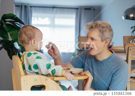 Young father feeding with a spoon adorable messy toddler baby sitting in high chair. Cute baby learning to eat food by himself. Kid enjoying healthy nutrition. family bonding moment during mealtime. Young father feeding with a spoon adorable messy toddler baby sitting in high chair. Cute baby learning to eat food by himself. Kid enjoying healthy nutrition. family bonding moment during mealtime. 135881166