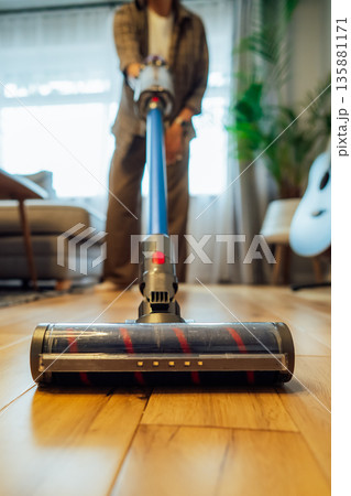 Focus on close up cordless vacuum cleaner vacuuming wooden floor of modern apartment living room with green plants. Daily routine and household chores. Housekeeping, home cleaning concept. Vertical. 135881171