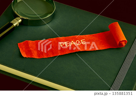 A red fabric scroll with the word Peace in gold letters rests on a dark green journal. A magnifying glass sits nearby, suggesting a close examination of the message 135881351