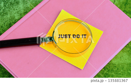 A magnifying glass focuses on the motivational phrase Just Do It, written on a yellow sticky note. The note rests on a pink book, which lies on a green textured surface A magnifying glass focuses on the motivational phrase Just Do It, written on a yellow sticky note. The note rests on a pink book, which lies on a green textured surface 135881367