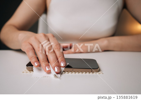 Close-up view of young woman cleaning smartphone screen with disinfectant wipe, preparing surface for installation of new glass screen protector, ensuring clean and bubble-free application. Close-up view of young woman cleaning smartphone screen with disinfectant wipe, preparing surface for installation of new glass screen protector, ensuring clean and bubble-free application. 135881619