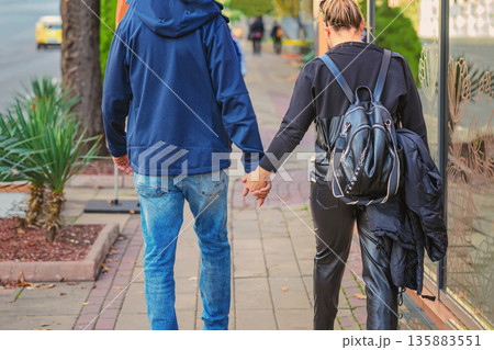 Loving couple walking hand in hand on a city street together, rear view. Back view of Couple walking hand in hand on urban sidewalk in casual clothes. Concept of love, connection,friendship Loving couple walking hand in hand on a city street together, rear view. Back view of Couple walking hand in hand on urban sidewalk in casual clothes. Concept of love, connection,friendship 135883551