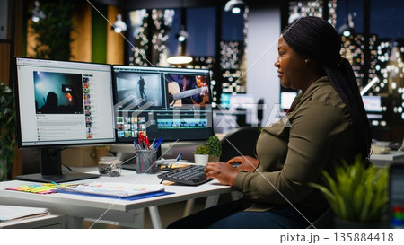 African american female videographer editing a film montage project, multitasking with dual monitors and professional software. Video editor in creative process for movie production. Camera B. 135884418