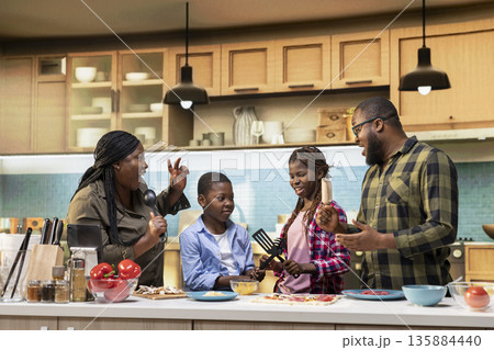 African American family enjoying silly karaoke session for pizza day in the kitchen, singing with utensils and doing goofy dance moves together at home. Weekend activity and cooking time. African American family enjoying silly karaoke session for pizza day in the kitchen, singing with utensils and doing goofy dance moves together at home. Weekend activity and cooking time. 135884440