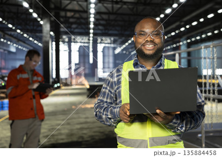 African american supervisor standing near steel machinery with laptop during construction tasks, showcasing production responsibility and the power of fabrication. Hi vis vest for safety. 135884458