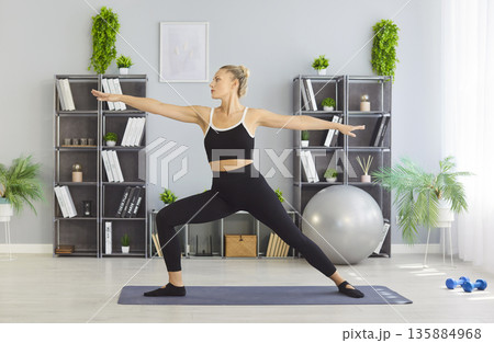 Woman practicing yoga warrior pose at home on mat for fitness training session Woman practicing yoga warrior pose at home on mat for fitness training session 135884968