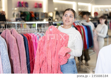 Girl stopped near leisure wear showcase, holds eiderdown bathrobe hanger in hands, examines product 135886184