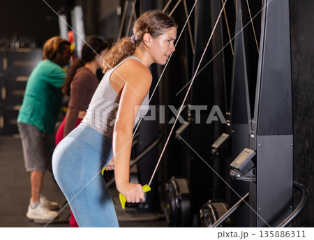Determined young girl training her arms using cable exercise machine in gym 135886311
