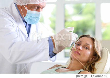 Male doctor gives injection and performs procedure of lip augmentation of female patient in clinic. Male doctor gives injection and performs procedure of lip augmentation of female patient in clinic. 135886440