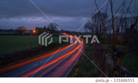 A peaceful countryside with a charming historic farm in the distance contrasted by the streaks of car light trails passing through the fields 135886555