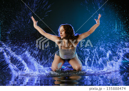 Female contemporary dancer kneels in shallow water creating splash in blue studio light. Use for website banner or advertising about dance classes, fitness, or performing arts. Female contemporary dancer kneels in shallow water creating splash in blue studio light. Use for website banner or advertising about dance classes, fitness, or performing arts. 135888474