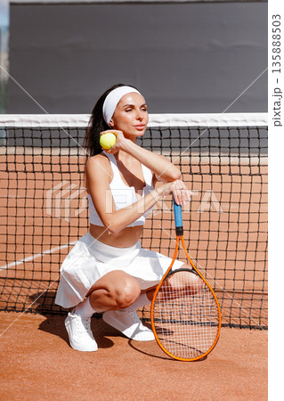 Female tennis player squats by the net holding ball and racket on clay court. Ideal for website banner or social ad about tennis training, fitness, and sport lifestyle. Female tennis player squats by the net holding ball and racket on clay court. Ideal for website banner or social ad about tennis training, fitness, and sport lifestyle. 135888503