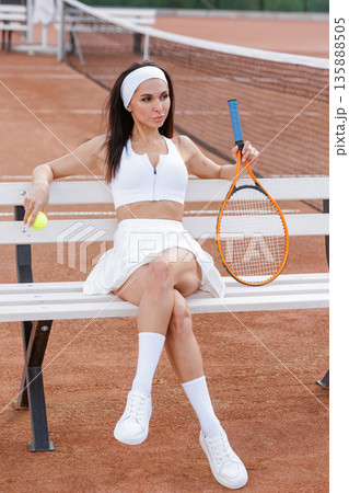 Tennis player rests on court bench holding ball beside racket. Ideal for website banner or social ad about training breaks, recovery, and sport lifestyle. Tennis player rests on court bench holding ball beside racket. Ideal for website banner or social ad about training breaks, recovery, and sport lifestyle. 135888505