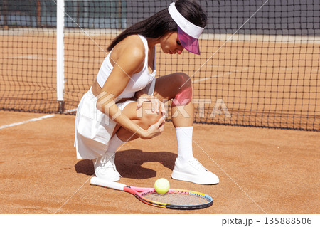 Athletic woman tennis player kneeling with knee pain on outdoor clay court, racket and ball nearby. Ideal for sports medicine, injury prevention, and training content on websites or ads. 135888506