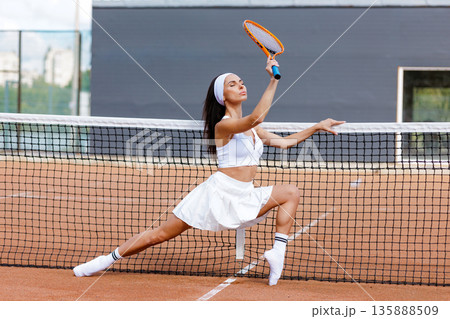 Athletic tennis player performs lunge stretch at the net on clay court. Ideal for website banner or social ad about tennis training, fitness, and warmup. Athletic tennis player performs lunge stretch at the net on clay court. Ideal for website banner or social ad about tennis training, fitness, and warmup. 135888509