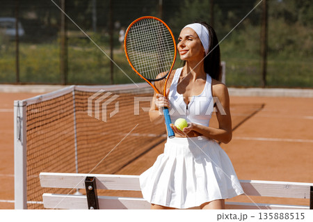 Female tennis player holds racket and ball near the net on clay court. Ideal for website banner or social ad about tennis training, fitness, and sport lifestyle. Female tennis player holds racket and ball near the net on clay court. Ideal for website banner or social ad about tennis training, fitness, and sport lifestyle. 135888515