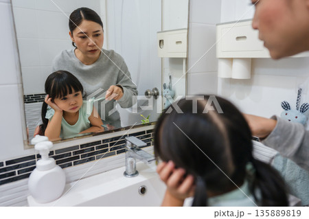stubborn child girl refusing to brush her teeth in the bathroom. mother is trying to encourage her unhappy daughter during the morning routine, while child looks grumpy and resistant. 135889819
