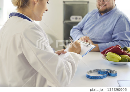 Nutritionist consulting overweight patient with vegetables, tape measure in clinic 135890906
