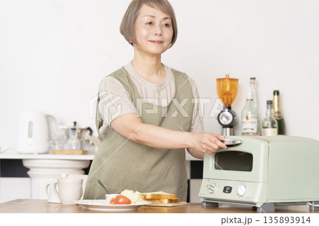 キッチンでトーストを焼いて朝食を食べるシニア女性 135893914