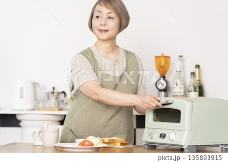 キッチンでトーストを焼いて朝食を食べるシニア女性 135893915