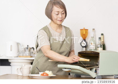 キッチンでトーストを焼いて朝食を食べるシニア女性 キッチンでトーストを焼いて朝食を食べるシニア女性 135893916