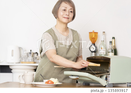 キッチンでトーストを焼いて朝食を食べるシニア女性 135893917