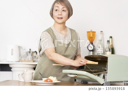 キッチンでトーストを焼いて朝食を食べるシニア女性 135893918