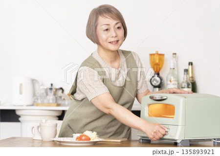 キッチンでトーストを焼いて朝食を食べるシニア女性 135893921