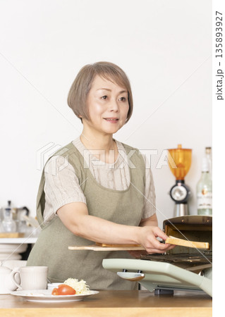 キッチンでトーストを焼いて朝食を食べるシニア女性 キッチンでトーストを焼いて朝食を食べるシニア女性 135893927
