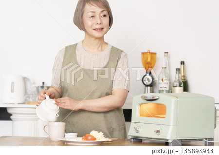 キッチンでトーストを焼いて朝食を食べるシニア女性 135893933