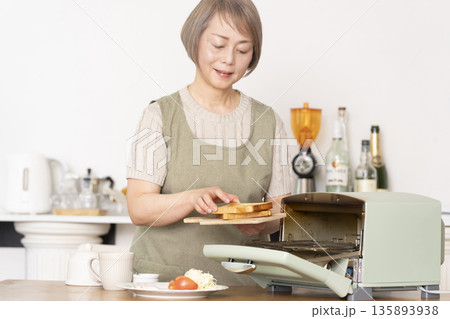 キッチンでトーストを焼いて朝食を食べるシニア女性 135893938