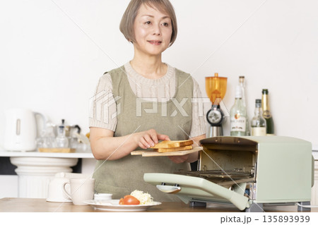 キッチンでトーストを焼いて朝食を食べるシニア女性 135893939