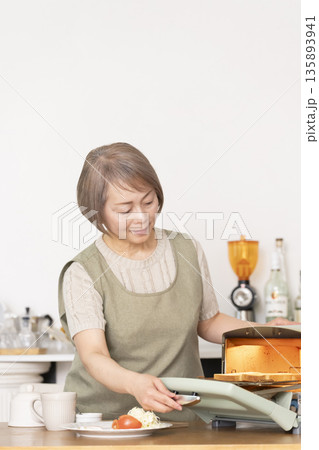 キッチンでトーストを焼いて朝食を食べるシニア女性 135893941