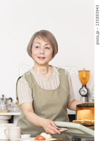 キッチンでトーストを焼いて朝食を食べるシニア女性 135893943