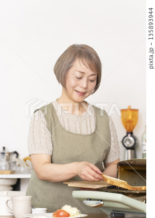 キッチンでトーストを焼いて朝食を食べるシニア女性 135893944